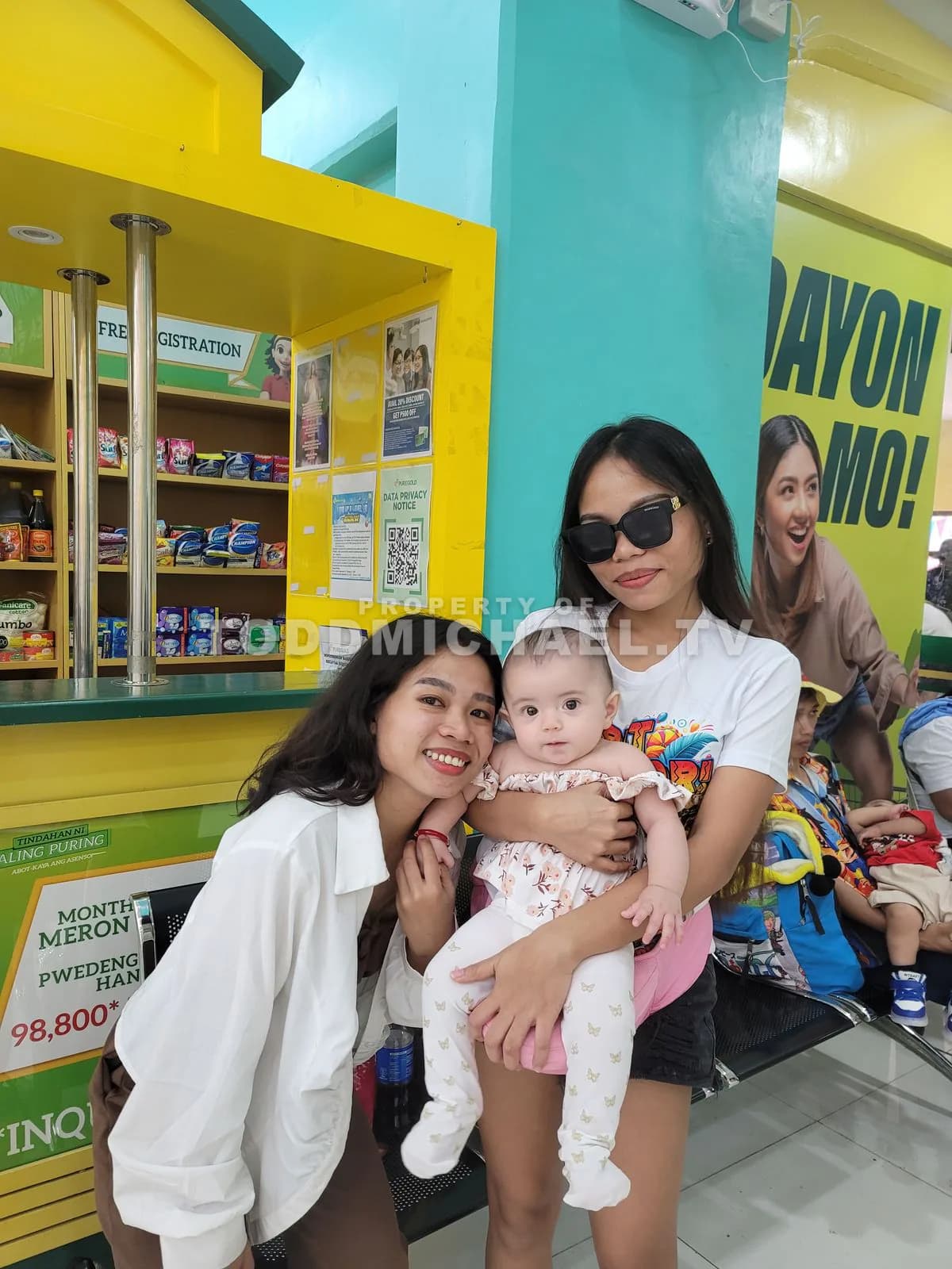 Two women and a baby in front of a brightly colored store with shelves filled with products.
