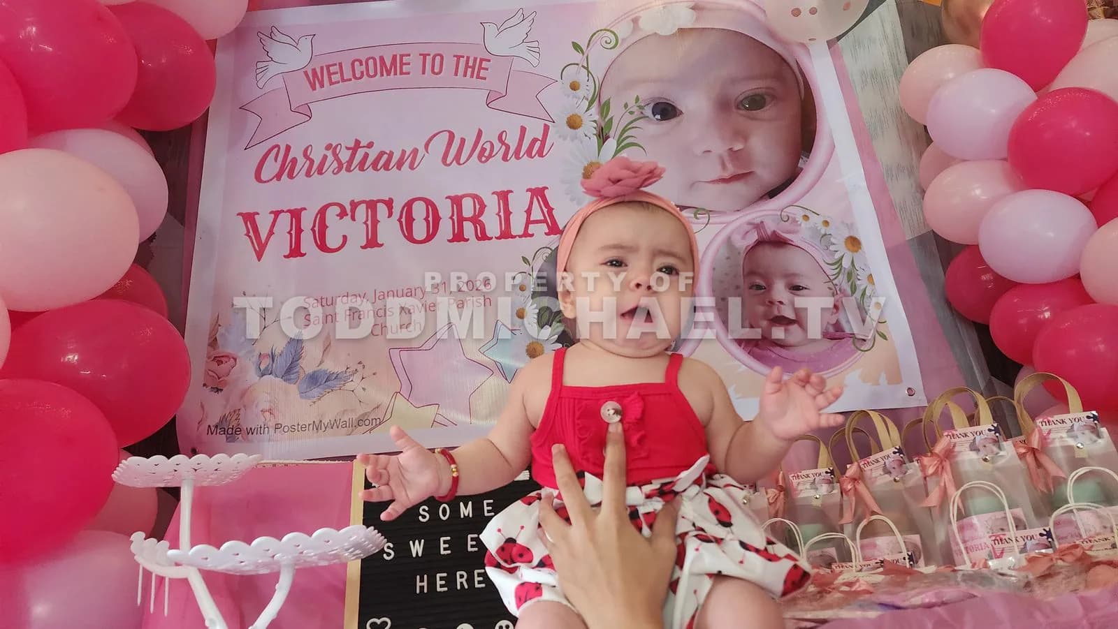 A baby sitting in front of a decorated table with balloons and a banner welcoming Victoria.