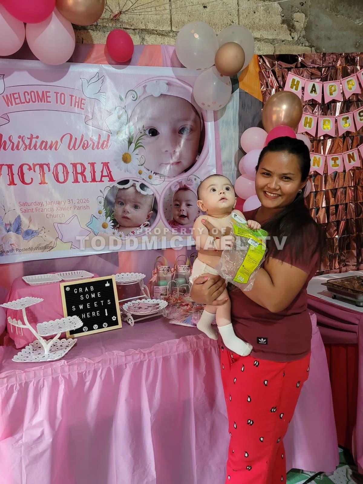 a woman holding a baby in front of a pink-themed party backdrop with balloons and decorations.