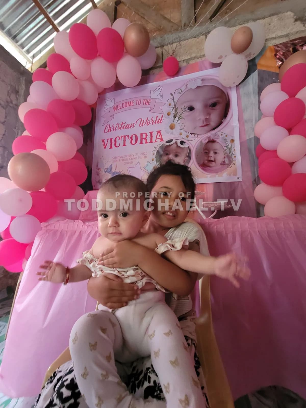 two children sitting on a chair with pink balloons and a banner celebrating Victoria's birthday.
