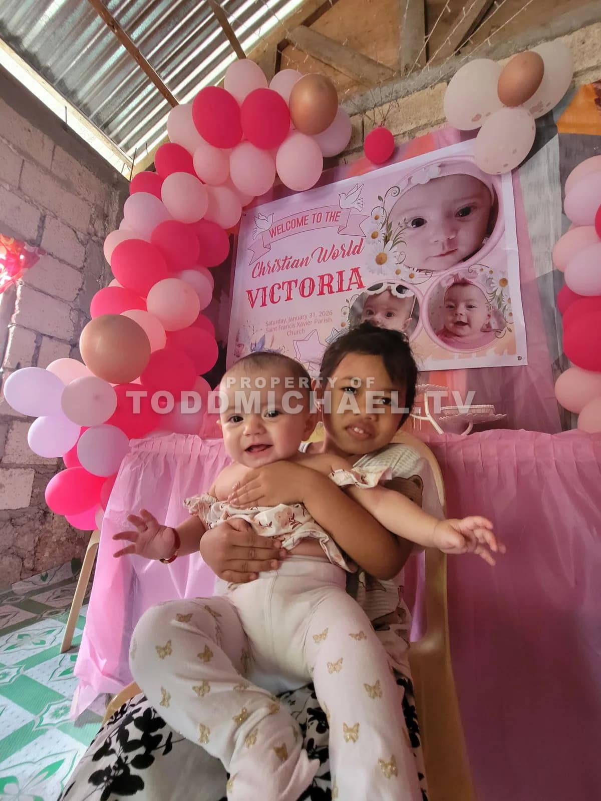 two children hugging in front of a balloon arch with 'Victoria' written on it.