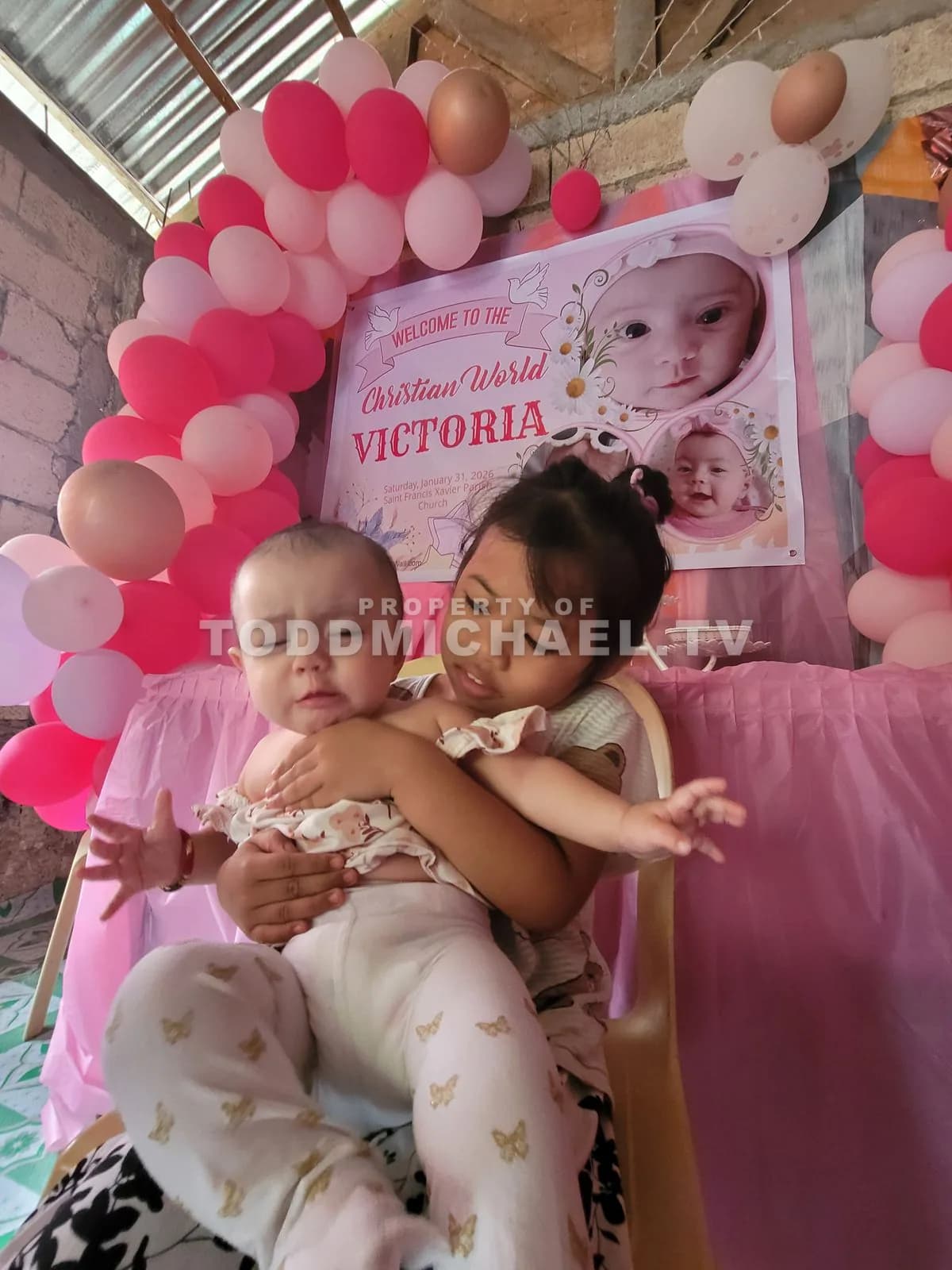 two children hugging in front of a balloon arch with 'Victoria' written on it.