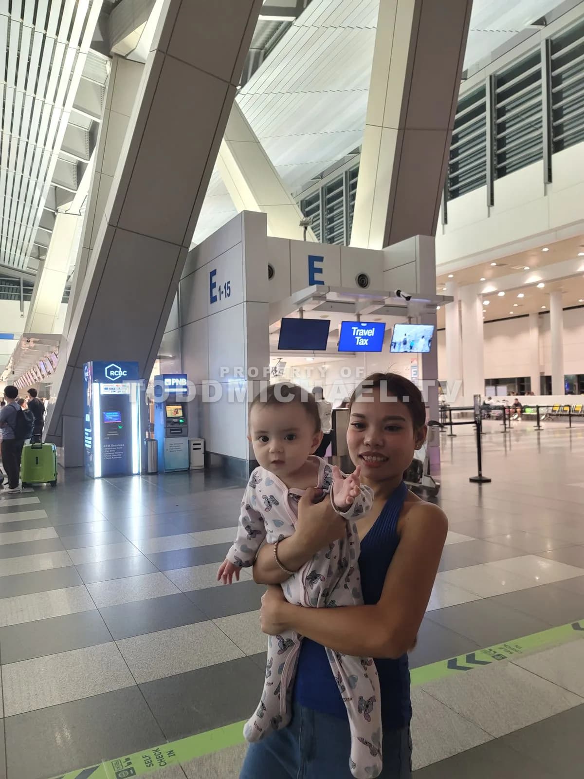 a woman holding a baby in an airport terminal with people and luggage around them.