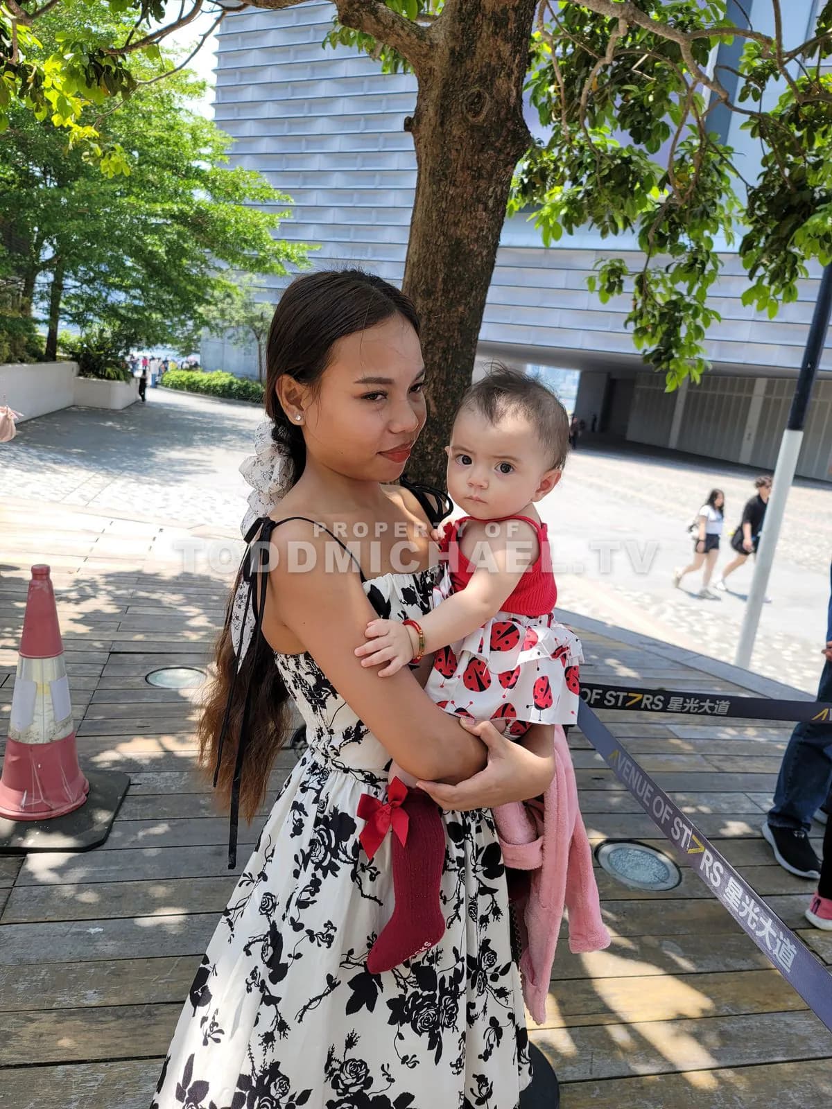 a woman holding a baby stands on a wooden platform with trees and Hong Kong's skyline in the background.