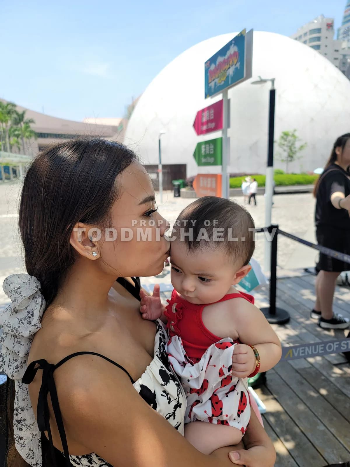 a woman kissing her baby while holding them in an outdoor setting with trees and modern architecture.